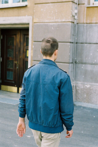 Eine Person mit kurzen Haaren, einer blauen Jacke und beigen Hosen, geht mit dem Rücken zur Kamera in der Nähe eines Gebäudeeingangs nach draußen.