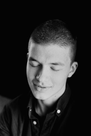Schwarz-Weiß-Porträt einer jungen Person mit kurzen Haaren, geschlossenen Augen und einem leichten Lächeln, die vor einem dunklen Hintergrund ein Hemd mit Knöpfen trägt.