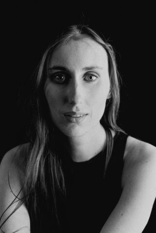 Schwarz-Weiß-Porträt einer Person mit langen Haaren, die ein ärmelloses Oberteil trägt und vor einem dunklen Hintergrund direkt in die Kamera blickt.