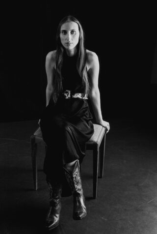 Eine Frau in einem ärmellosen Kleid und Cowboystiefeln sitzt vor einem dunklen Hintergrund auf einer Bank und blickt direkt in die Kamera.