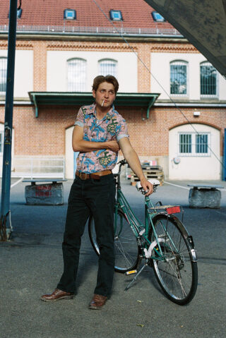 Ein Mann in einem gemusterten Hemd und einer dunklen Hose steht mit verschränkten Armen und einer Zigarette in der Hand neben einem grünen Fahrrad in einer städtischen Umgebung im Freien.