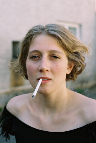 Draußen steht eine Person mit kurzen, welligen blonden Haaren und einem schwarzen schulterfreien Top und hält eine brennende Zigarette im Mund.