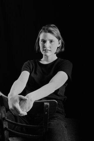 Eine Person mit kurzen Haaren sitzt rückwärts auf einem Stuhl, die Arme ruhen auf der Rückenlehne und blickt in die Kamera. Das Bild ist schwarz-weiß mit dunklem Hintergrund.