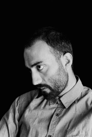 Schwarzweißfoto eines Mannes mit kurzen Haaren und Bart, der ein zugeknöpftes Hemd trägt und vor einem schwarzen Hintergrund zur Seite blickt.