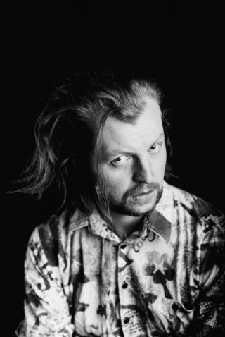 Ein Mann mit langen Haaren und Bart, der ein gemustertes Hemd trägt, blickt vor einem schwarzen Hintergrund in die Kamera.