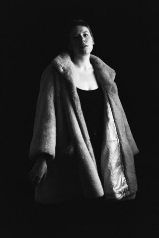 Vor einem dunklen Hintergrund steht eine Person, die einen hellen Pelzmantel über einem dunklen Outfit trägt, mit neutralem Gesichtsausdruck und leicht geneigtem Kopf.