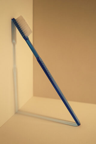 Eine blaue Zahnbürste steht aufrecht und lehnt mit ihren Borsten an der Ecke, wo zwei beige Wände aufeinandertreffen und wirft einen Schatten auf die Oberflächen.