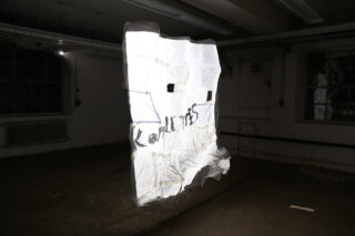 Ein weißes, durchscheinendes Blatt mit schwarzem Text hängt in einem dunklen, leeren Raum mit Industriefenstern und freiliegenden Rohren.