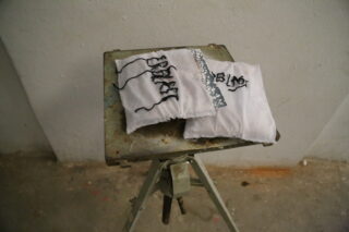 Auf einem kleinen Metallhocker stehen vor einem schlichten Wandhintergrund zwei weiße Stoffstücke mit schwarz gesticktem Text und Mustern, eines davon mit einem Streifen silbernen Klebebands.