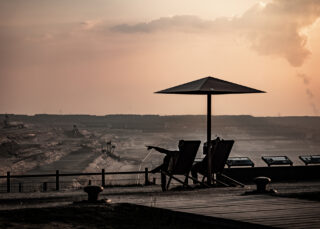 Zwei Personen sitzen auf Liegestühlen unter einem Sonnenschirm und blicken auf eine riesige industrielle Bergbaulandschaft bei Sonnenuntergang.
