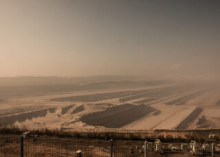 Unter einem dunstigen Himmel erstreckt sich in der Ferne ein breiter, staubiger Tagebau. Im Vordergrund sind hinter einem Zaun terrassierte Schichten und Maschinen zu sehen.