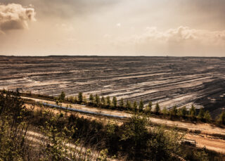 Ein großer Tagebau mit terrassierten Schichten und spärlicher Vegetation im Vordergrund unter einem dunstigen Himmel.