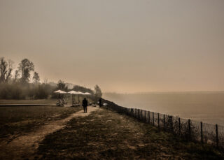 Eine Person geht unter einem dunstigen Himmel auf einem Feldweg in der Nähe eines eingezäunten Uferbereichs mit geschlossenen Sonnenschirmen und vereinzelten Bäumen entlang.