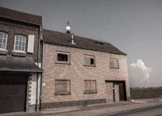 Ein vernageltes, verlassenes Backsteingebäude mit zerbrochenen Fenstern steht neben einem älteren Haus in einer verlassenen Straße unter klarem Himmel.