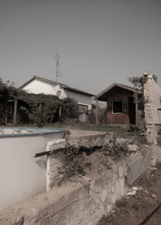 Eine beschädigte Betonmauer und ein leerer Pool vor zwei kleinen Häusern, mit überwucherter Vegetation und klarem Himmel im Hintergrund.