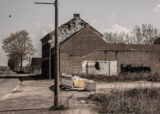 Neben einer verlassenen Straße steht ein verlassenes Gebäude mit vernagelten Fenstern und Graffiti. Auf dem Boden liegen einige Absperrungen und Trümmer.