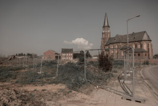 Eine eingezäunte Baustelle mit überwuchertem Gras neben einer Straße, mit einer großen alten Kirche und einigen Backsteingebäuden im Hintergrund unter einem klaren Himmel.