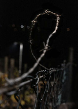 Nahaufnahme von Stacheldraht auf einem Maschendrahtzaun bei Nacht, mit unscharfen Hintergrundlichtern und sichtbaren Zaunpfosten.