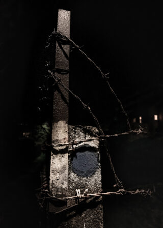 Ein Betonpfosten mit abblätternder Farbe wird von einer vertikalen Metallstange gekrönt und ist mit rostigem Stacheldraht umwickelt. Nachts vor einem dunklen Hintergrund fotografiert.