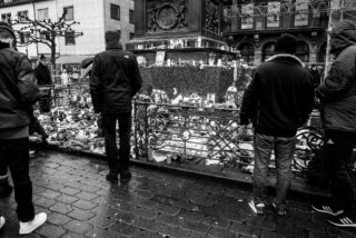 Schwarzweißfoto von Menschen, die in einer Stadtstraße neben einem provisorischen Denkmal mit Blumen, Kerzen und Fotos stehen.