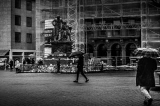 An einem regnerischen Tag gehen Menschen mit Regenschirmen über einen Stadtplatz und kommen dabei an einer von Blumen umgebenen Statue und einem im Bau befindlichen Gebäude im Hintergrund vorbei.