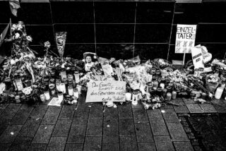 Ein Denkmal mit Kerzen, Blumen, Fotos und handgeschriebenen Schildern in deutscher Sprache, angeordnet auf einem Bürgersteig vor einer gefliesten Wand.