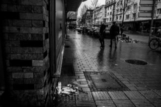 Eine verregnete Stadtstraße mit verstreutem Müll auf dem Bürgersteig und zwei Menschen, die in der Ferne spazieren gehen; Gebäude und geparkte Autos säumen die Straße.