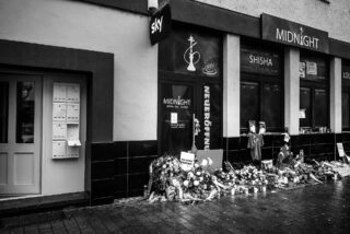 Vor einer Shisha-Bar und nahegelegenen Geschäften in einer Stadtstraße ist ein Denkmal mit Blumen, Kerzen und Notizen aufgestellt.