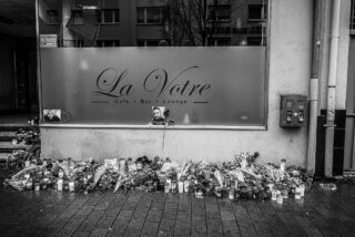 Vor dem Eingang eines Cafés namens „La Votre“ ist auf einem verregneten Gehweg ein Denkmal mit Blumen, Kerzen und Fotos aufgestellt.