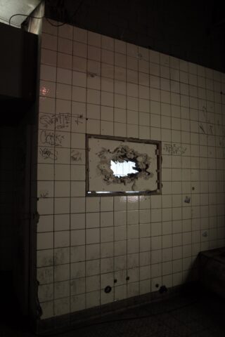 Eine Wand aus weißen Fliesen mit Graffiti und einem großen Loch in der Mitte, das den Blick auf ein kleines Fenster und etwas Tageslicht freigibt.