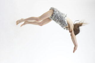 Eine Person in einem hellgrauen Rüschenoutfit scheint vor einem schlichten weißen Hintergrund in der Luft zu schweben oder zu fallen, die Arme nach vorne ausgestreckt und das Haar weht.