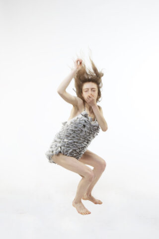 Eine Person mit langen Haaren und einem strukturierten Kleid ist vor einem weißen Hintergrund in der Luft gefangen, mit einer Hand in der Nähe des Mundes und der anderen erhoben.