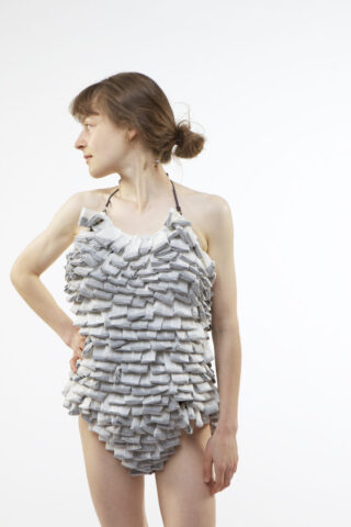 Eine Frau steht in einem strukturierten, grauen Badeanzug mit Rüschen vor einem schlichten weißen Hintergrund und blickt mit einer Hand auf der Hüfte nach links.