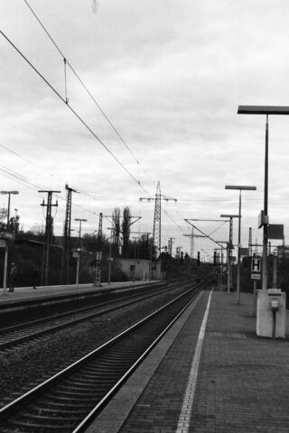 Schwarzweißfoto eines leeren Bahnsteigs mit mehreren Gleisen, Oberleitungen und bewölktem Himmel.