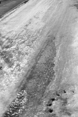 Eine schneebedeckte Straße mit einem sichtbaren Streifen geräumten Asphalts und Reifenspuren, die schmelzenden Schnee und Eisflecken zeigen.