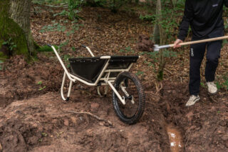 Eine Person schaufelt auf unebenem, schlammigem Boden in einem Waldgebiet Erde in eine Schubkarre.