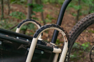 Nahaufnahme des Griffs und Hinterrads eines schlammigen Fahrradanhängers mit Waldboden und grünem Laub im Hintergrund.