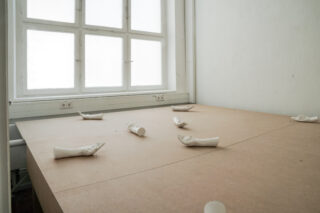 Ein einfacher Tisch mit sieben auf seiner Oberfläche angeordneten weißen Handskulpturen in einem minimalistisch eingerichteten Raum mit einem großen Fenster im Hintergrund.