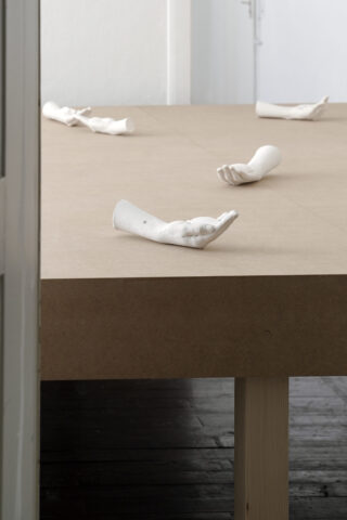 In einem minimalistischen Raum mit weißen Wänden sind mehrere weiße Gipsabdrücke von Händen auf einem einfachen Holztisch angeordnet.