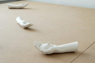 Drei weiße Gipsabdrücke von Unterarmen und Händen sind auf einer einfachen Holzoberfläche angeordnet, jede Hand ist offen und nach oben gerichtet.