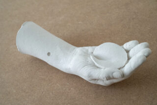 Eine weiße Gipsskulptur eines menschlichen Unterarms und einer Hand hält ein flaches, rundes Objekt, das auf einer braunen, strukturierten Oberfläche präsentiert wird.