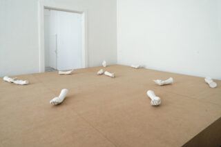 In einem minimalistischen Raum mit weißen Wänden und einer offenen Tür sind mehrere weiße Gipsabdrücke menschlicher Arme und Hände auf einer großen Holzfläche angeordnet.