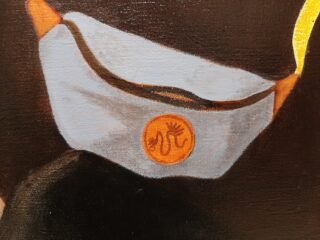 Eine Nahaufnahme einer grauen Gesichtsmaske mit orangefarbenen Ohrschlaufen und einem runden orangefarbenen Emblem mit einem schlangenartigen Design auf der Vorderseite.