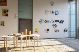 Drei Holztische mit Kunstbedarf und Holzklötzen stehen in einem sonnendurchfluteten Raum. An den Wänden hängen abstrakte Kunstwerke, und durch die Fenster fällt natürliches Licht herein.