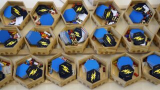 Sechseckige Holztabletts enthalten blaue und schwarze sechseckige Karten, farbige Pillen und Zettel, die in Reihen auf einer flachen Oberfläche angeordnet sind.