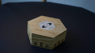 Auf einer dunklen Oberfläche steht eine sechseckige Holzbox mit einem Etikett mit einer schwarzweißen Grafik und Text.