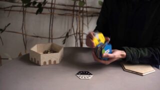 Eine Person mischt an einem Tisch mit einem sechseckigen Holzbehälter und einer schwarz-weißen sechseckigen Karte ein buntes Kartenspiel.