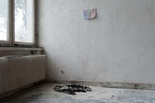 Ein fast leerer Raum mit einem Heizkörper, einem großen Fenster, einer kleinen bunten Tapete an der Wand und einem geschwärzten Pulverfleck auf dem Boden in der Nähe der Ecke.