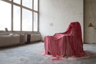 Ein Stuhl in einem leeren Raum ist mit einem rot gefärbten Stoff bedeckt, der sich auf dem Boden verteilt, neben großen Fenstern und einem Heizkörper.