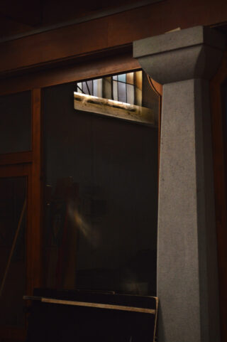 Ein schwach beleuchteter Innenraum mit einer großen grauen Säule, Glasplatten mit Holzrahmen und einem kleinen Fenster in der Nähe der Decke, das natürliches Licht hereinlässt.